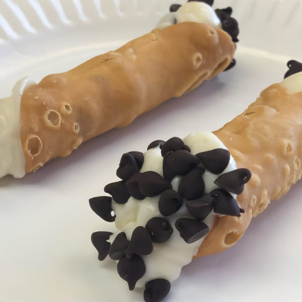 Two J&M Italian American Cuisine cannolis, one topped with chocolate chips, on a white plate.